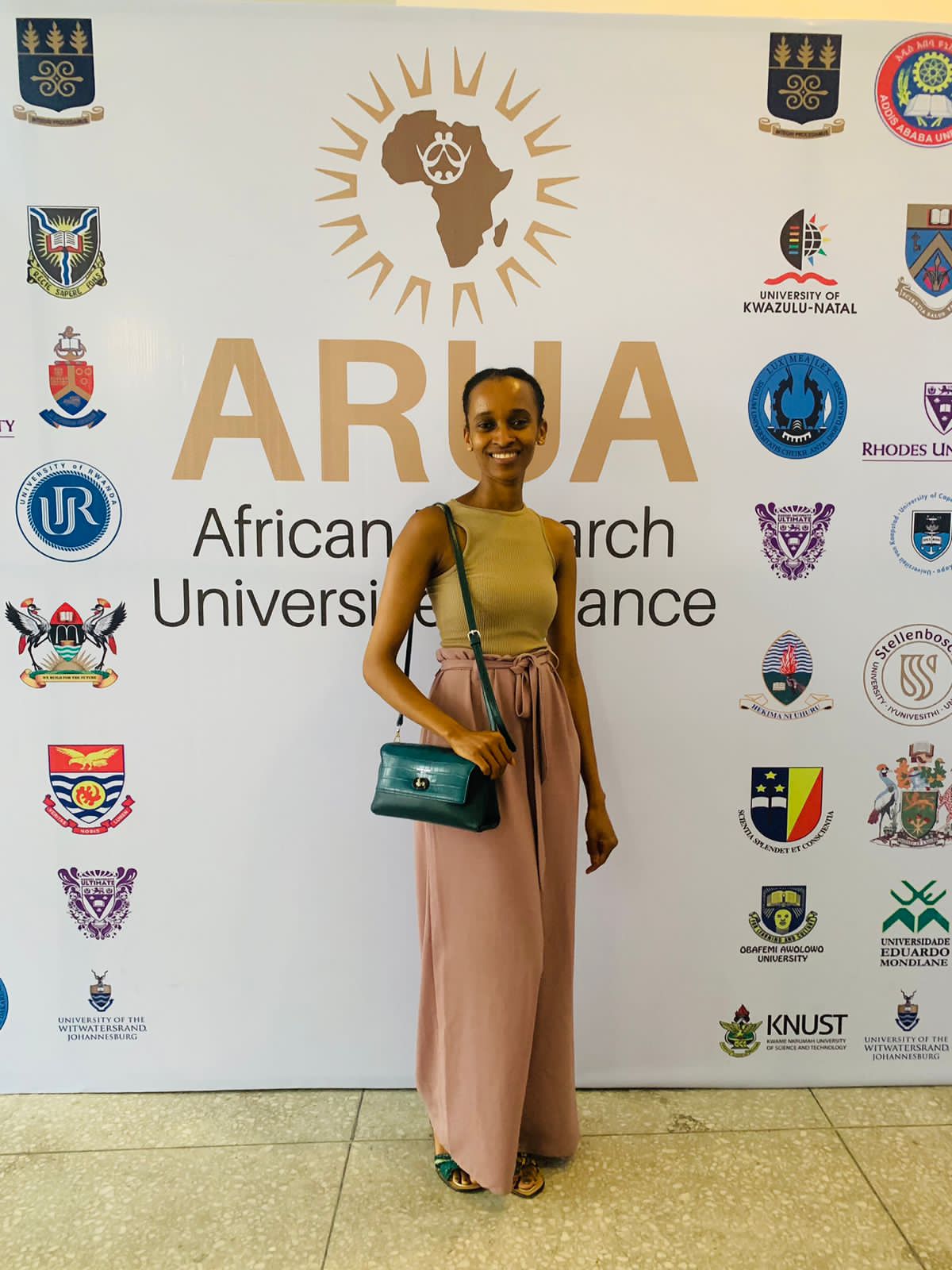 Standing in front of a backdrop for the African Research Universities Alliance, wearing wide-leg trousers and a crossbody bag.