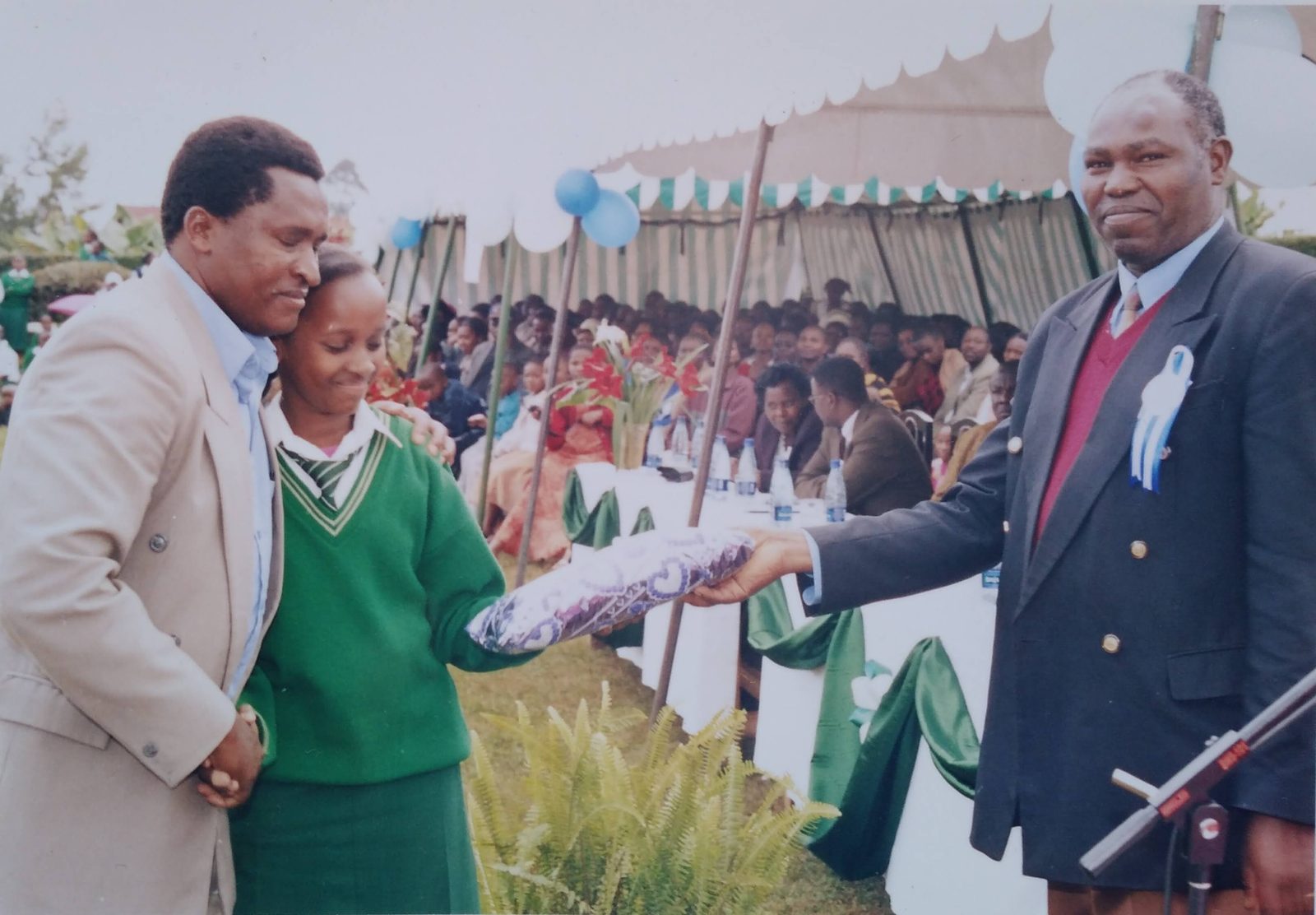 A schoolgirl in a green uniform is congratulated as she receives a wrapped prize at an outdoor school ceremony.
