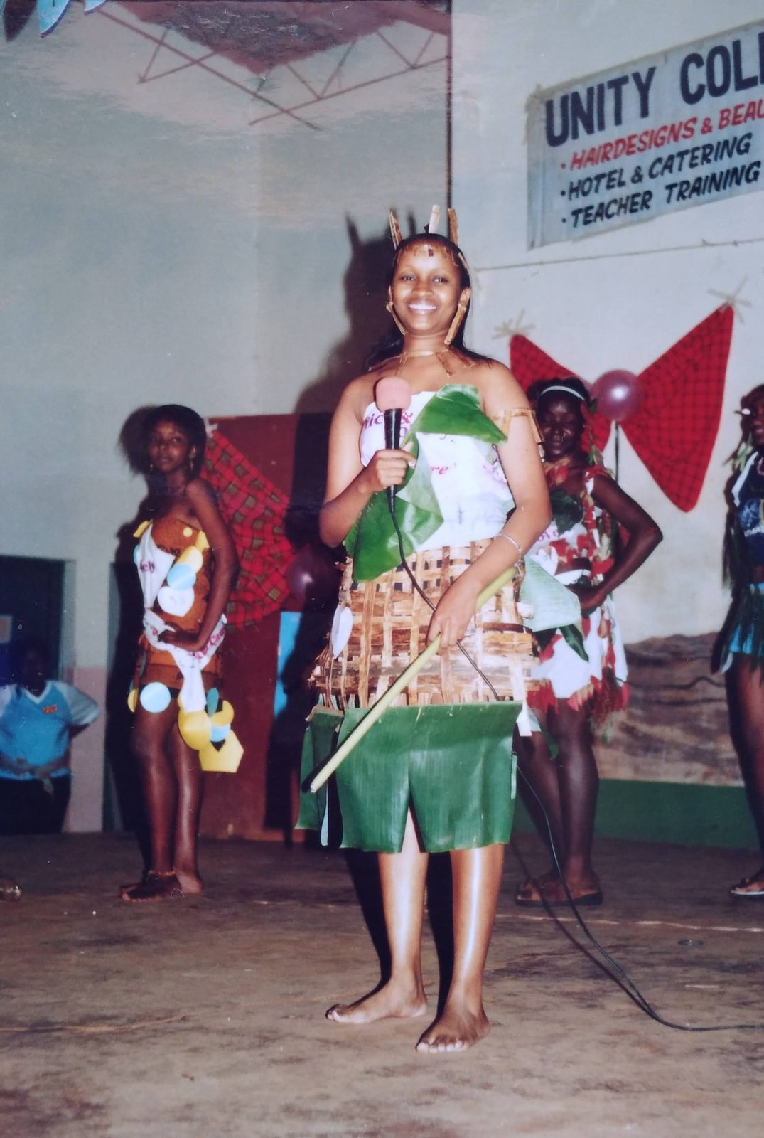 A student on stage holding a microphone in a costume made from banana leaves and woven natural fibres, with other models behind her.