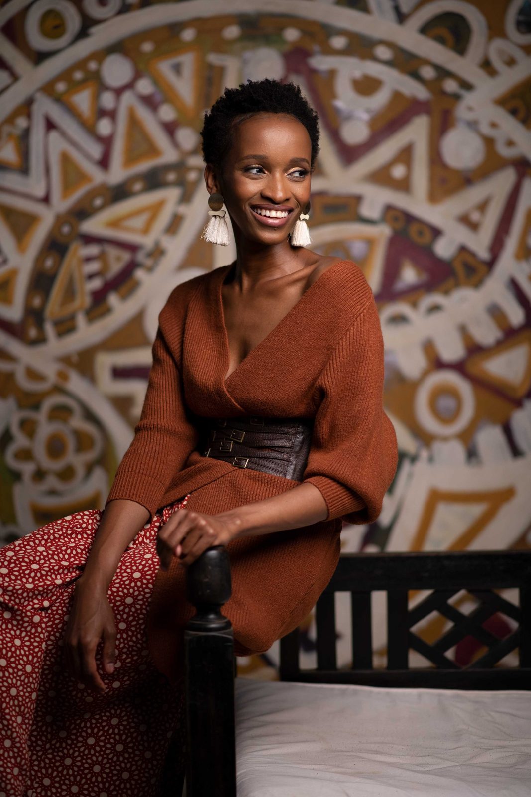 A model in a rust knit wrap top, corset belt and patterned skirt, seated against a painted mural.