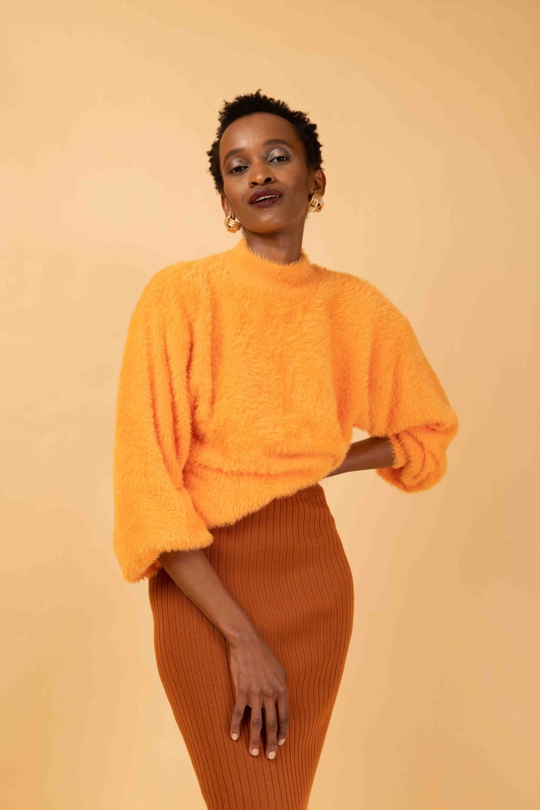 A model in a fluffy orange batwing sweater and rust ribbed skirt against a warm studio backdrop.
